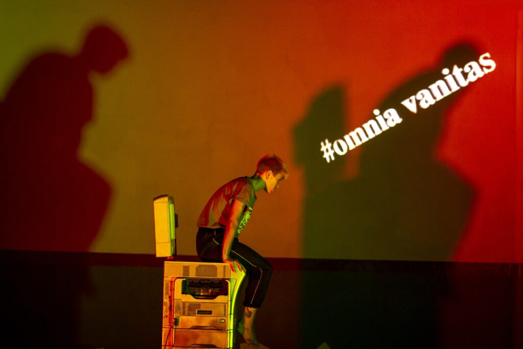 Performer sitting on a printer in Omnia Vanitas by Salon de los Invisibles